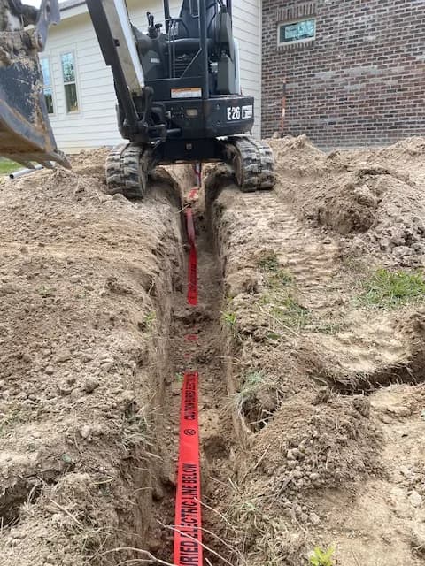 Bobcat E26 mini-excavator actively trenching with red CAUTION ELECTRIC LINE BELOW tape laid in the trench